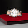 The TUDOR 1926 wristwatch features a silver and rose gold bracelet and white dial, showcased against a red and black surface with a textured black background.