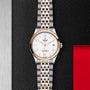 A TUDOR 1926 wristwatch with a silver and rose gold band displayed on a textured black surface.