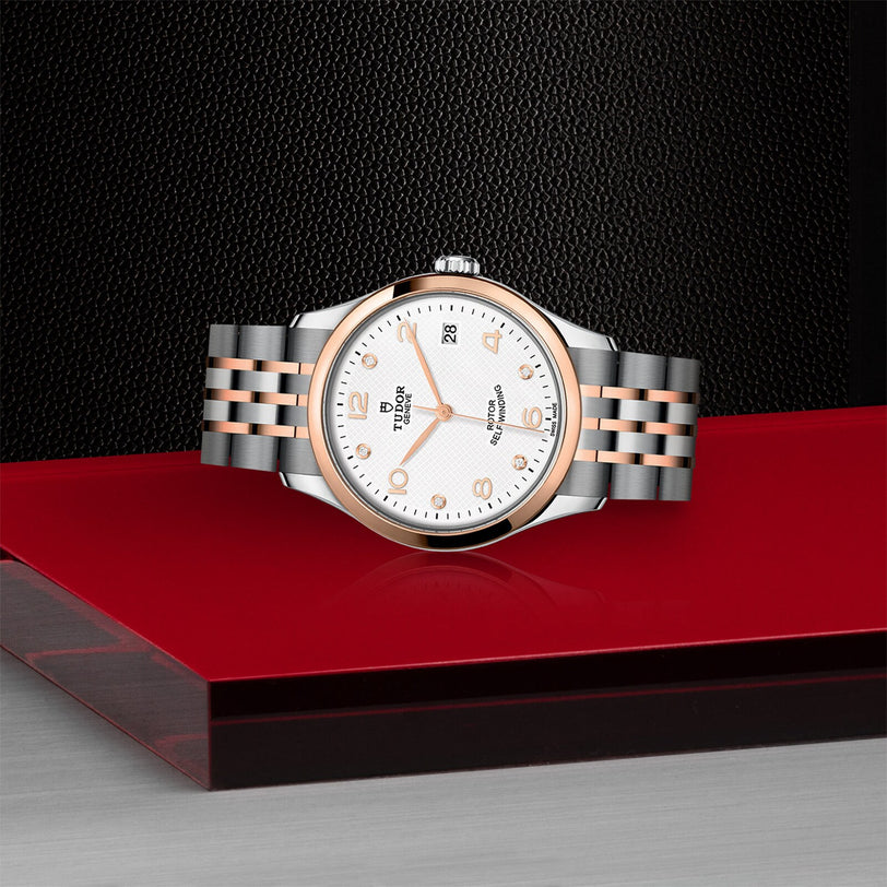 The TUDOR 1926 wristwatch features a white dial, gold accents, and a metal bracelet—an elegant two-tone timepiece displayed on red and black, perfect for those who appreciate classic style.