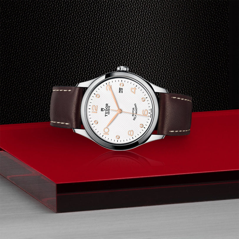 The TUDOR 1926 wristwatch, with its elegant dial, gold hour markers, and brown leather strap, rests on a red surface against a textured black background—a timeless piece inspired by classic mechanical watches.