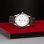The TUDOR 1926 wristwatch, with its elegant dial, gold hour markers, and brown leather strap, rests on a red surface against a textured black background—a timeless piece inspired by classic mechanical watches.
