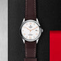 A TUDOR 1926 wristwatch with a brown leather strap and white dial displayed on a textured black and red background.