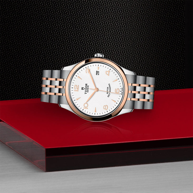 The TUDOR 1926 wristwatch, featuring a silver and rose gold metal band, is displayed on a glossy red and black surface against a textured black backdrop—ideal for fans of finely crafted mechanical timepieces.