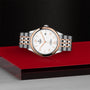 The TUDOR 1926 wristwatch, featuring a silver and rose gold metal band, is displayed on a glossy red and black surface against a textured black backdrop—ideal for fans of finely crafted mechanical timepieces.