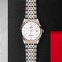 The TUDOR 1926 wristwatch, featuring a silver and rose gold band, white dial with gold markers and hands, and date display, rests on a textured black surface with a red and silver backdrop—showcasing classic elegance.