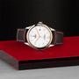 A TUDOR 1926 watch with a white dial, brown leather strap, and silver case is showcased on a red and black geometric surface against a textured black background, capturing the timeless elegance of mechanical timepieces.