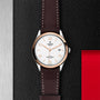 A TUDOR 1926 watch with a white dial, gold accents, brown leather strap, and date display sits on a textured black surface beside metal and red details—a classic choice for mechanical watch enthusiasts.