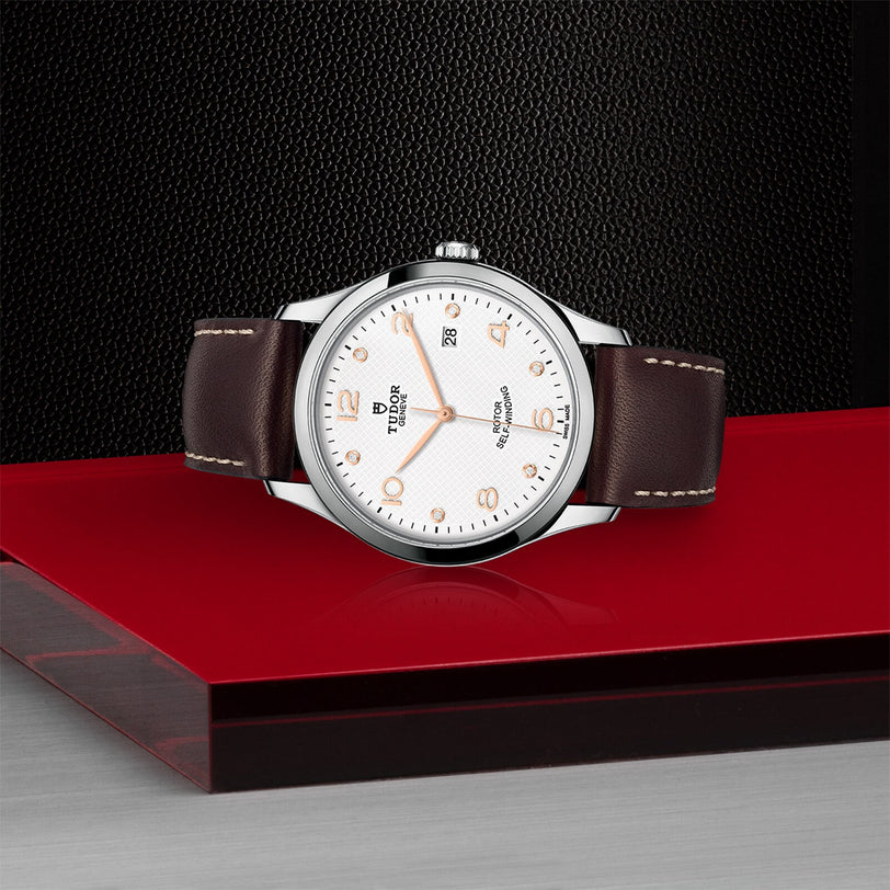 The TUDOR 1926 wristwatch, featuring a white dial, gold markers, and a brown leather strap, is showcased on a red and black surface with a textured black background.
