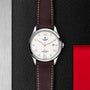 The TUDOR 1926 features a white dial, brown leather strap, silver case, gold-tone indices, and date display. Resting on a textured black surface with red and silver accents, its a timeless mechanical watch for classic style lovers.