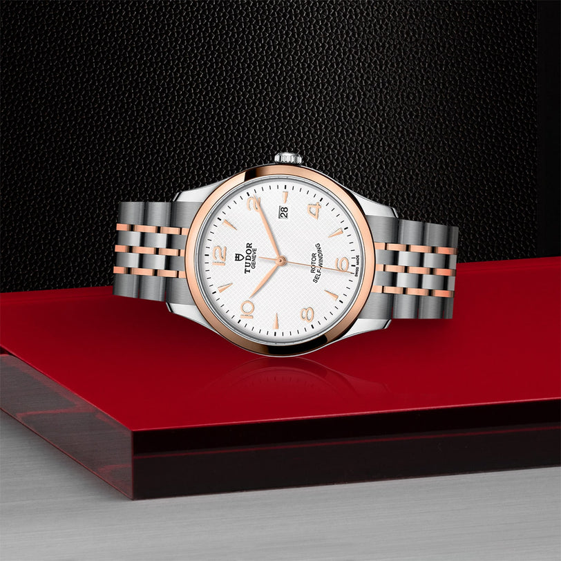 A TUDOR 1926 wristwatch with a silver and rose gold metal band, white dial, and date display rests elegantly on a glossy red and black surface—a refined homage to classic mechanical timepieces.