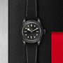 TUDOR Black Bay Ceramic wristwatch with a black strap, displayed on a textured black and red background.