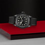 The TUDOR Black Bay Ceramic wristwatch, featuring a black face with white hour markers, is showcased on a sleek black and red platform, highlighting the Swiss-made watchs timeless elegance against a textured backdrop.
