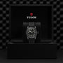 A Swiss-made TUDOR Black Bay Ceramic wristwatch with a black strap is displayed in an open black box, featuring the TUDOR logo and branding on the inside of the lid.