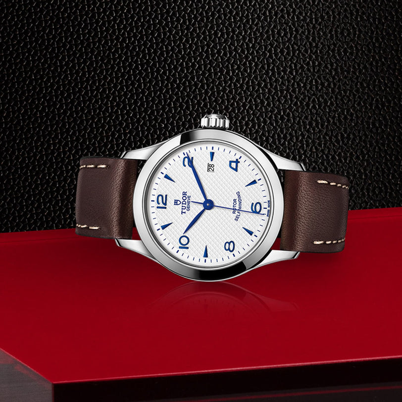 A TUDOR 1926 wristwatch with a white textured dial, blue numerals and hands, and a brown leather strap is displayed on a red surface against a black textured background.