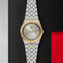 A TUDOR Royal wristwatch in silver and gold, featuring a grey dial with date display, displayed on a textured black surface.