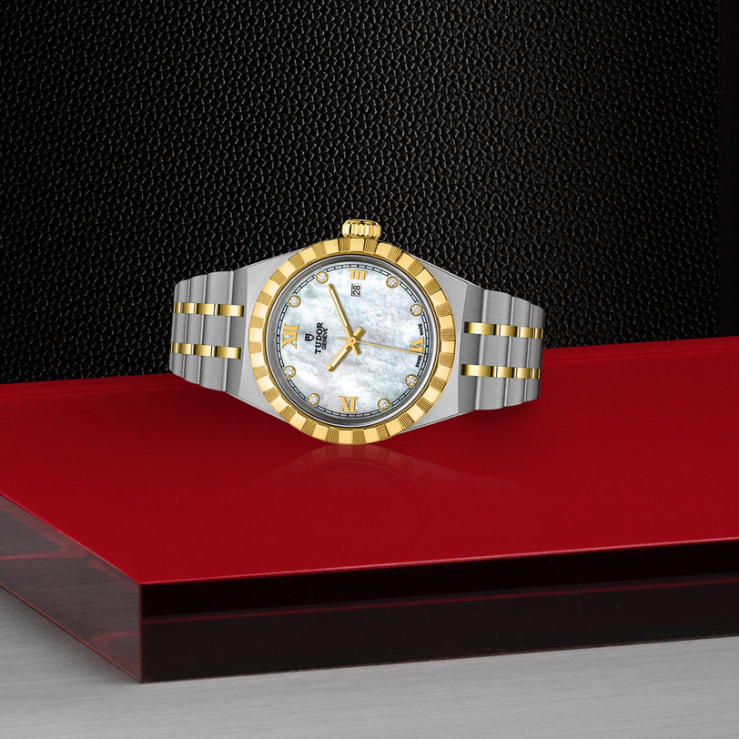 The TUDOR Royal luxury wristwatch, featuring a silver and gold design with a white face, is displayed on a red and black textured surface.