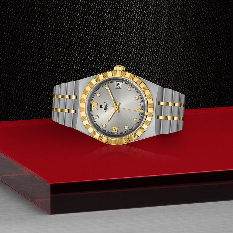 A TUDOR Royal two-tone gold and silver wristwatch displayed on a red and black textured surface.