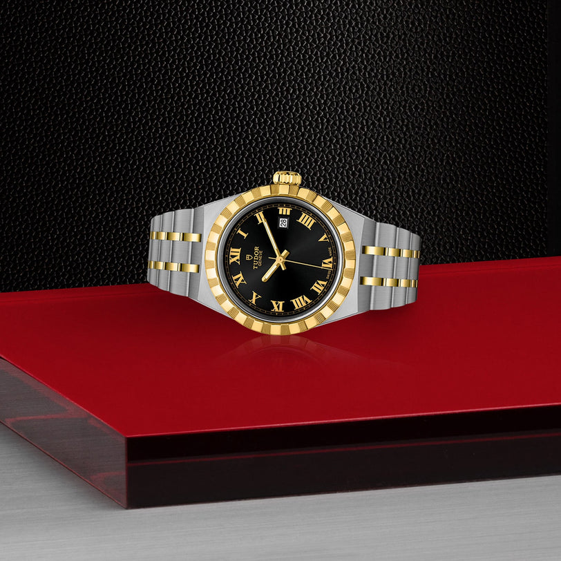 A TUDOR Royal watch with a black dial, gold Roman numerals, and a gold bezel in stainless steel and gold rests on a red and silver surface against a textured black background.