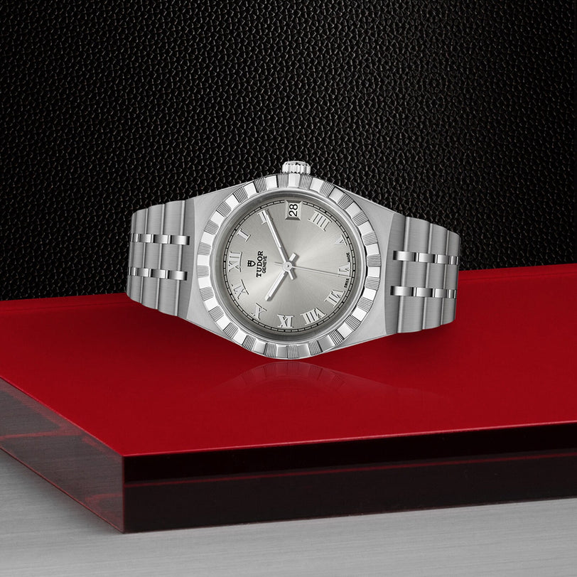 A TUDOR Royal wristwatch with a stainless steel and gold metallic bracelet and Roman numeral hour markers is shown on a red and black surface against a textured black background.