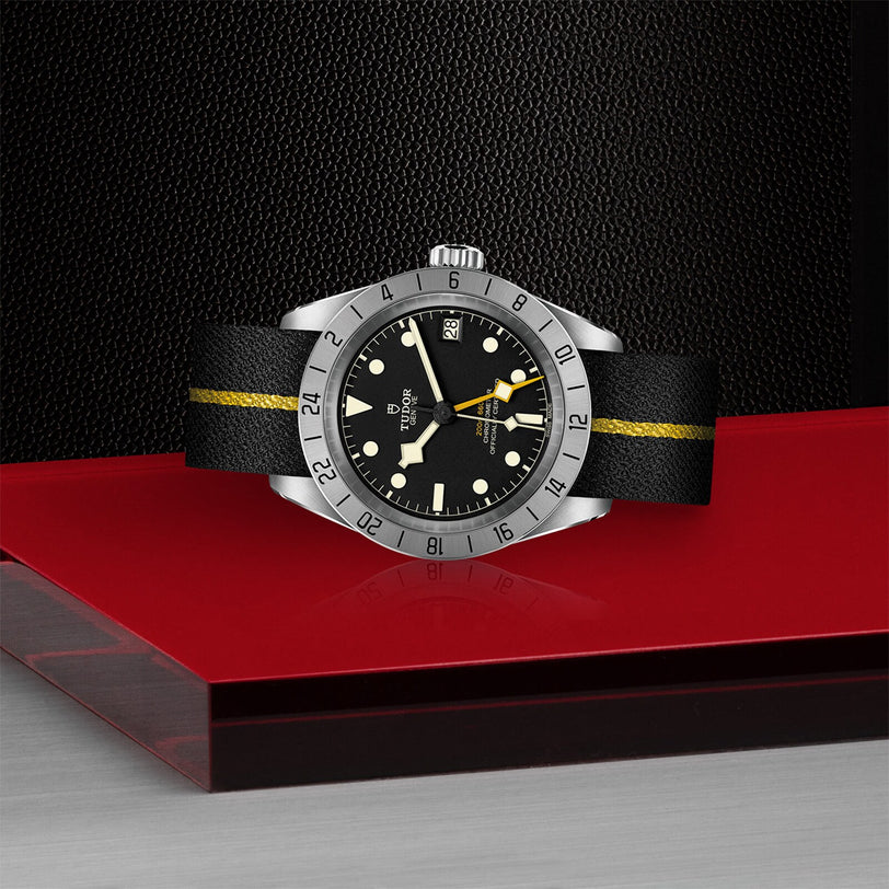 The TUDOR Black Bay Pro, a neo-vintage wristwatch with a black dial, silver bezel, and black strap with yellow stripe, is showcased on a red and metallic surface against a textured black background.
