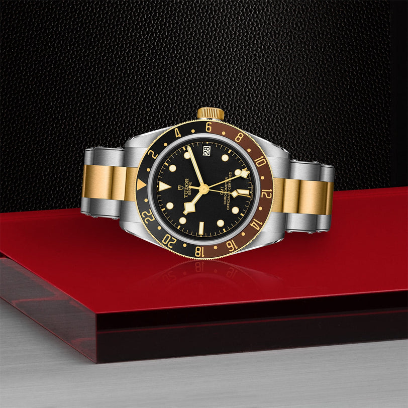 The TUDOR Black Bay GMT S&G wristwatch, featuring yellow gold accents and a black dial and bezel, is showcased on a red and gray platform against a textured black background.
