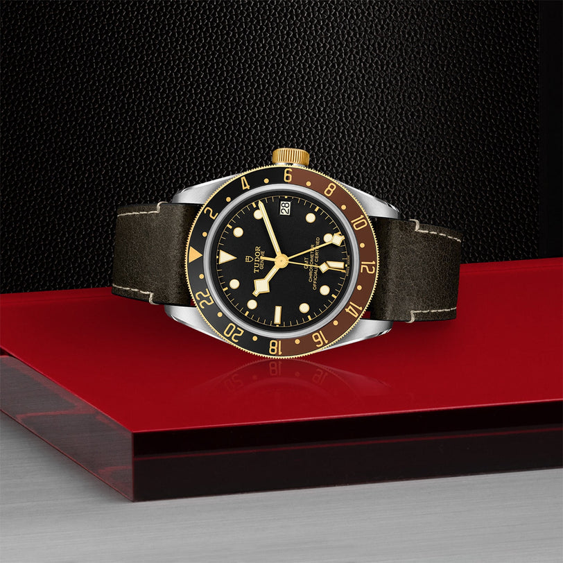 The TUDOR Black Bay GMT S&G, a black and yellow gold wristwatch with a leather strap inspired by Black Bay divers, is displayed on a red and gray surface against a textured black background.