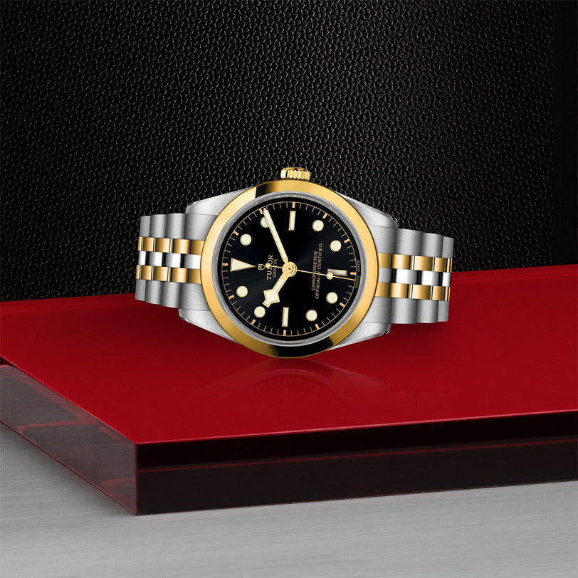 The TUDOR Black Bay One 36 S&G, featuring a black dial, gold bezel, and silver-and-gold bracelet, is displayed on a red and silver surface against a black textured background.