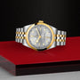 The TUDOR Black Bay One 36 S&G, a two-tone wristwatch with a silver face, gold bezel, and metal link strap, is displayed on a red surface against a textured black background.