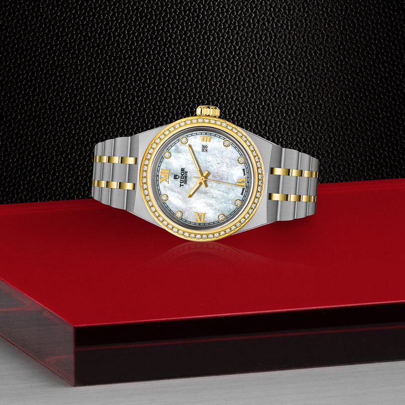 The TUDOR Royal, a luxury two-tone wristwatch with a diamond bezel and mother-of-pearl dial, is showcased on a red platform against a textured black background.