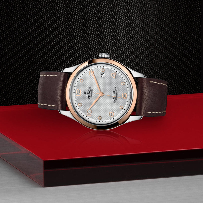 A TUDOR 1926 wristwatch featuring a brown leather strap, silver dial, gold accents, and date display is displayed on a red and gray surface with a black textured background.