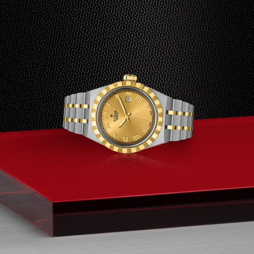 A TUDOR Royal wristwatch in two-tone gold and stainless steel, featuring a gold dial with Roman numerals, is displayed on a red and black geometric surface against a textured black background.