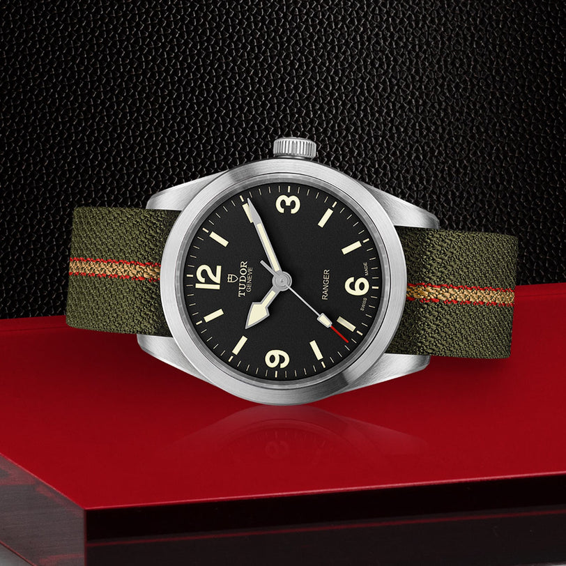 The TUDOR Ranger wristwatch, featuring a black dial with white numerals and an olive green jacquard strap, is shown on a red surface against a textured black background. It’s powered by the TUDOR MT5402 movement.