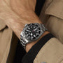 A person in a tan jacket wears the TUDOR Pelagos 39, a stainless steel wristwatch with a black dial and silver bracelet.