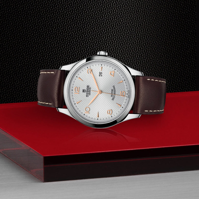 A TUDOR 1926 watch with a brown leather strap and white dial is displayed on a red and gray platform against a textured black background, representing the legacy of TUDOR.