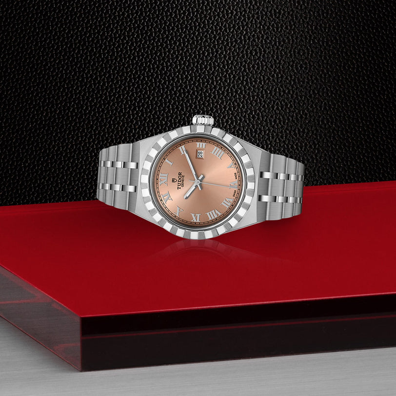 A TUDOR Royal wristwatch with a salmon dial and Roman numerals is displayed on a glossy red and black geometric surface, exemplifying effortless sport-chic style.