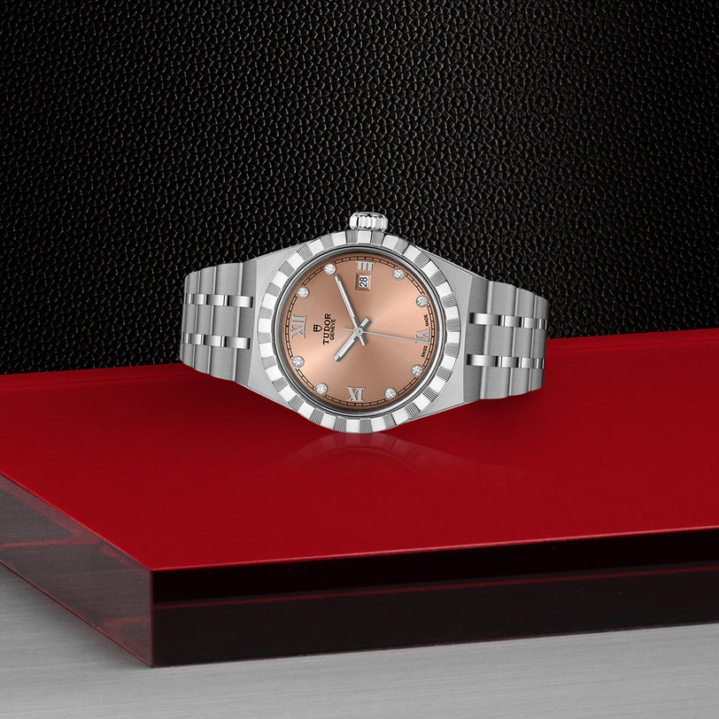 A TUDOR Royal wristwatch with a salmon dial and Roman numerals, featuring automatic movement, is displayed on a red and black geometric surface with a textured black backdrop.