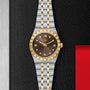 A TUDOR Royal watch featuring a gold and silver metal band, gold bezel, brown dial with date display, automatic movement, and diamond hour markers is displayed on a textured black surface for a sporty chic look.
