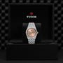 A TUDOR Royal wristwatch with a silver band and salmon dial, featuring automatic movement, is displayed upright in an open black presentation box branded with the TUDOR logo.