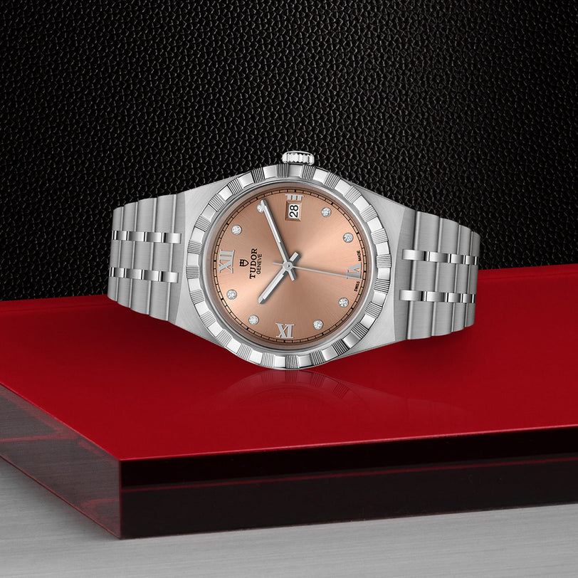 A TUDOR Royal stainless steel wristwatch with a rose gold dial, diamond markers, and a diamond-set bezel rests on a glossy red and black surface against a textured black background.