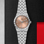 A silver TUDOR Royal wristwatch with a salmon dial, Roman numerals, diamond markers, date display, and automatic movement rests on a textured black surface near a red edge.