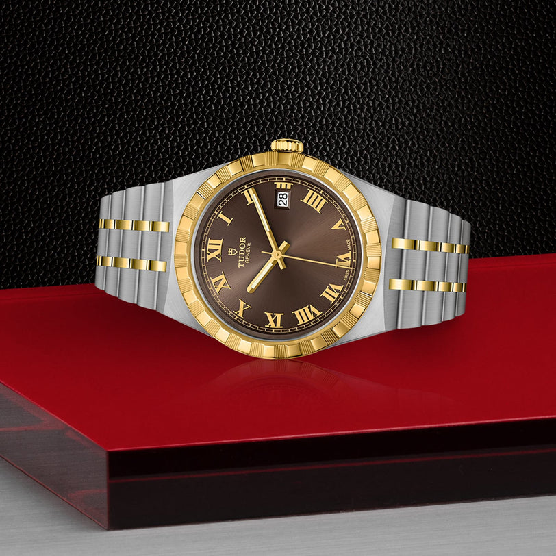 The TUDOR Royal wristwatch features a gold and silver band, a brown face with Roman numerals, and is shown on a red and black surface.