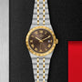 A sport-chic TUDOR Royal wristwatch with a gold and silver metal band, gold bezel, black dial with Roman numerals and date display, set against a textured black and metallic backdrop. Features reliable automatic movement.