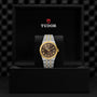 A TUDOR Royal wristwatch with a gold and silver band and a black and gold dial, featuring automatic movement, is showcased in an open black Tudor-branded box.