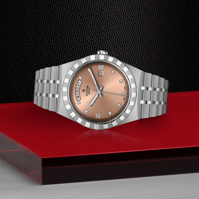 A TUDOR Royal wristwatch with a salmon dial, diamond-set bezel, and day-date display is showcased on a red and black geometric surface, blending sport-chic style with elegance.