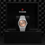A TUDOR Royal wristwatch with a brown gradient dial, diamond-set bezel, and metal bracelet is displayed upright in a black box with the Tudor logo, highlighting the sophisticated sport-chic style of the TUDOR Royal.