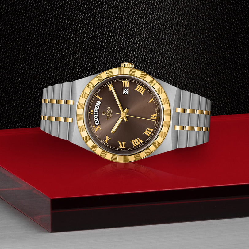 A chic TUDOR Royal wristwatch with a gold bezel, brown dial, Roman numerals, day-date display, and automatic movement is displayed on a red and black surface against a textured black background.
