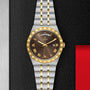 A TUDOR Royal wristwatch with a gold and silver metal band, brown dial, gold Roman numerals, and day-date display—featuring automatic movement—rests on a textured black surface with a red accent.