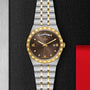 The TUDOR Royal is a sporty chic wristwatch featuring a gold and silver band, black dial with gold bezel, Roman numerals, diamond markers, automatic movement, and day-date displays on a textured black surface.