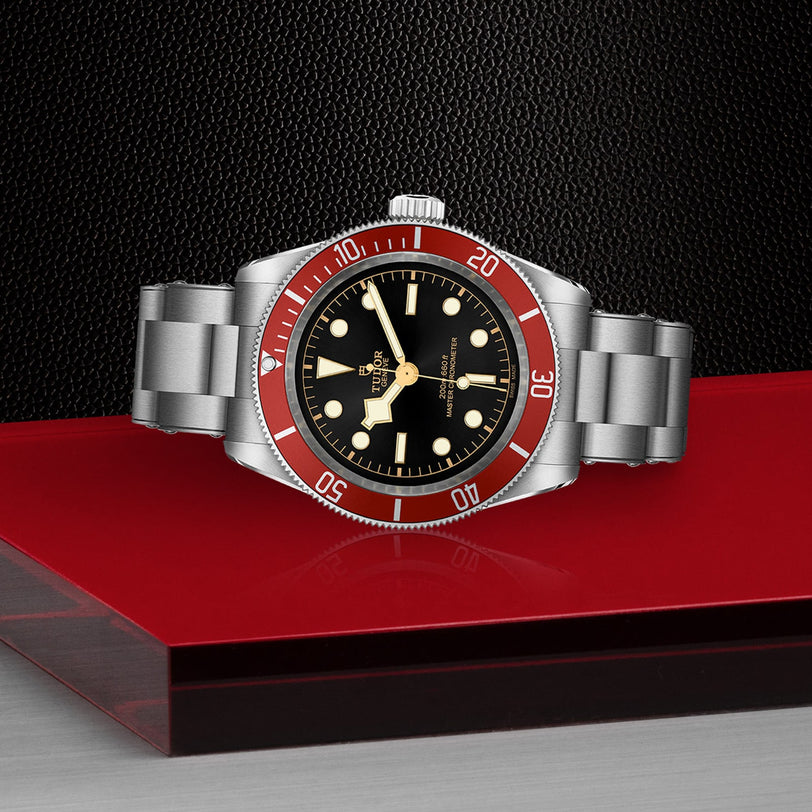 A TUDOR Black Bay stainless steel wristwatch with a black dial and burgundy bezel, showing 10:11, rests on a glossy red and black surface against a textured black background.