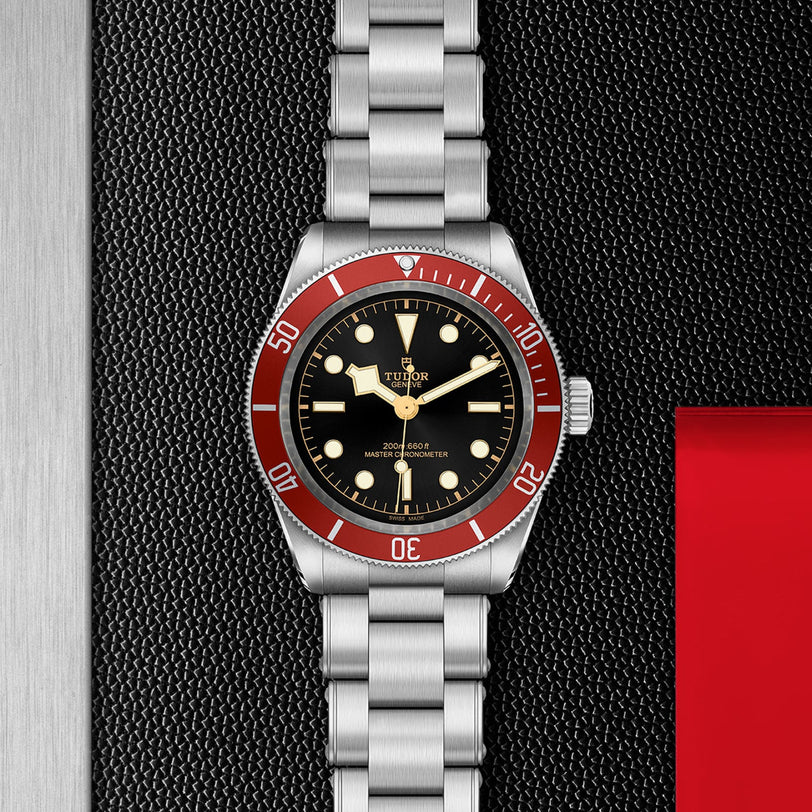 A TUDOR Black Bay wristwatch with a stainless steel bracelet, black dial, and bold burgundy bezel is showcased against a textured black and metallic backdrop.